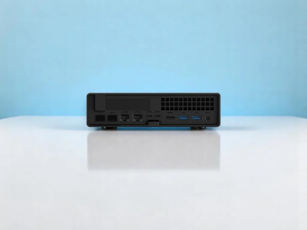 MinisForum MS-A2. Вид сзади компактного черного мини-ПК с множеством портов, включая USB, LAN, SFP+, HDMI и питание, на блестящем белом столе с голубым фоном