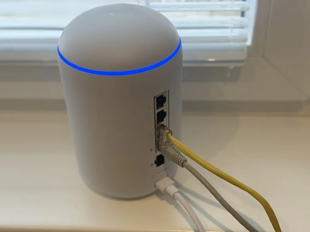 UniFi Dream Router 7. Современный белый цилиндрический Wi-Fi роутер с синей светодиодной подсветкой и несколькими подключёнными Ethernet кабелями, установленный на белой поверхности у окна с жалюзи
