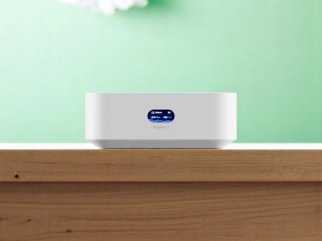 Белый WiFi маршрутизатор UniFi Express 7 с цифровым дисплеем, показывающим количество клиентов и скорость сети на деревянной поверхности на зелёном фоне