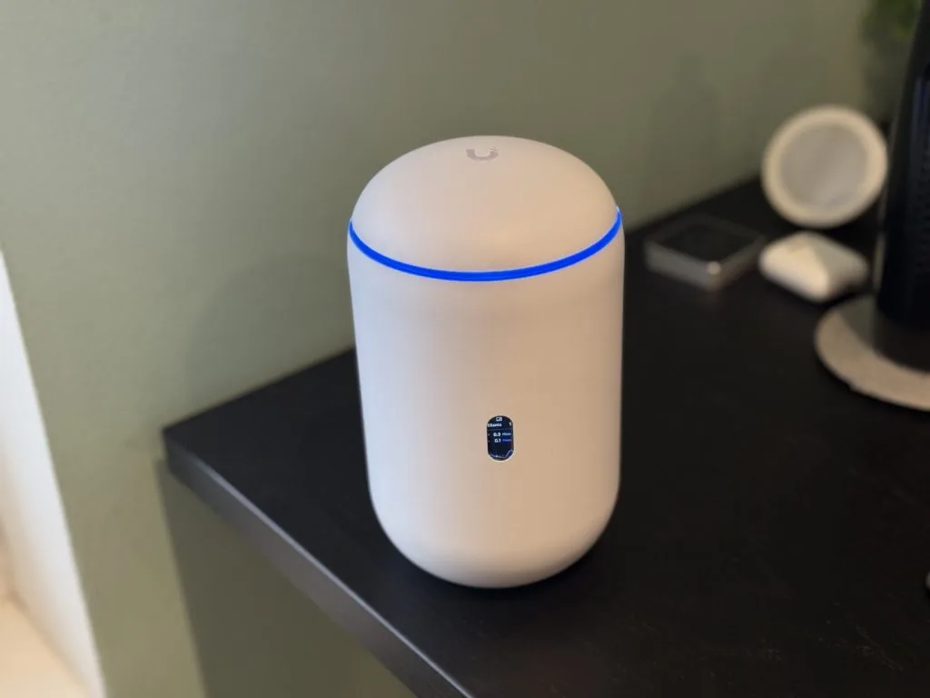 UDR7, Белый Wi-Fi роутер Ubiquiti с синим светодиодным кольцом на черном столе рядом с другими устройствами